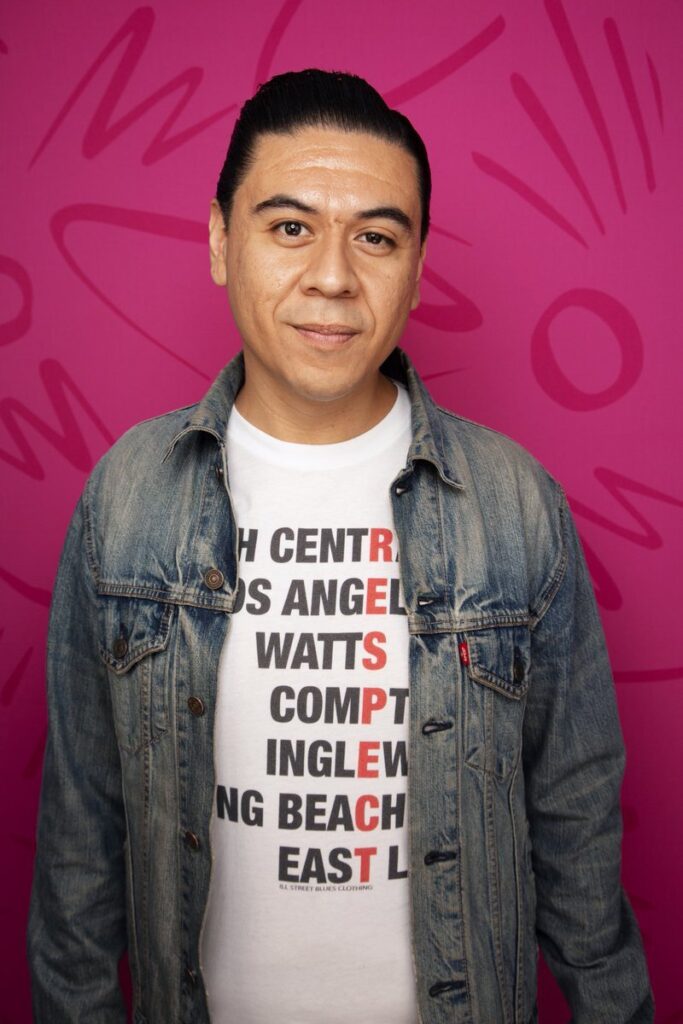 Photo of Chris Estrada in a Los Angeles-themed tee shirt. He's standing in front of a pink background. Photo taken by Mindy Tucker.