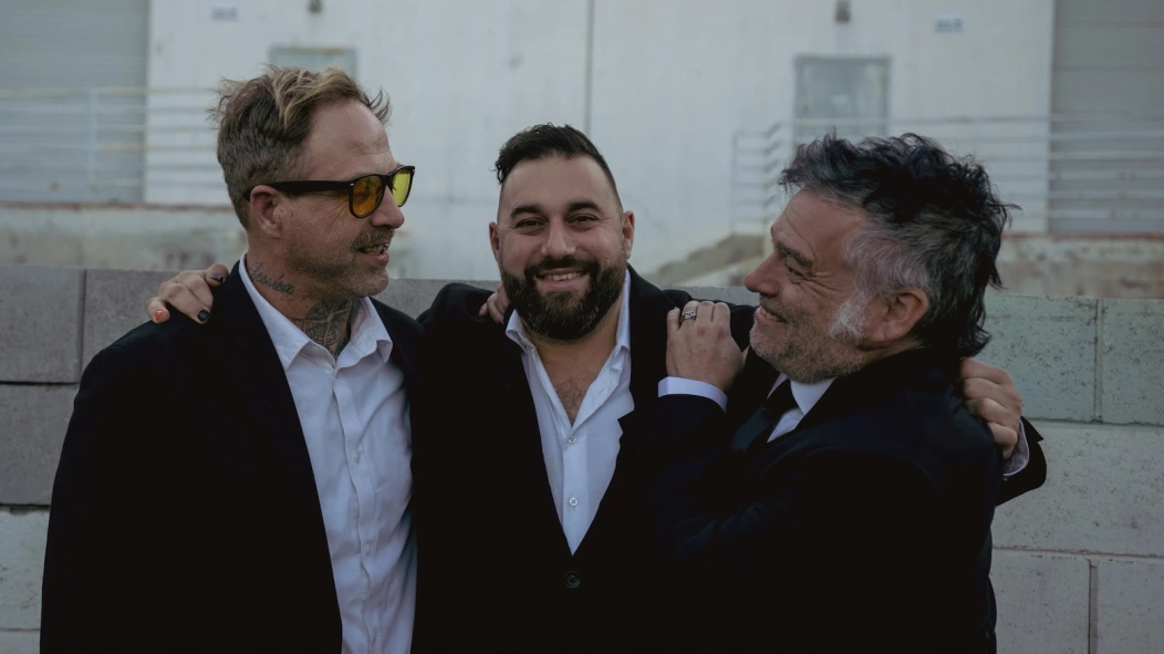 Sam King, Ceschi Ramos and Fat Mike hugging on a rooftop. All wearing white button-down shirts and black suit jackets.